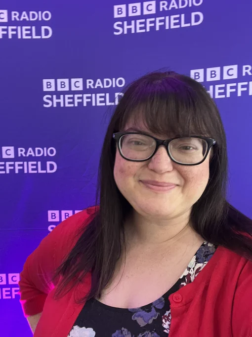Chella poses in front of the bbc radio Sheffield logo in a floral dress and red cardigan