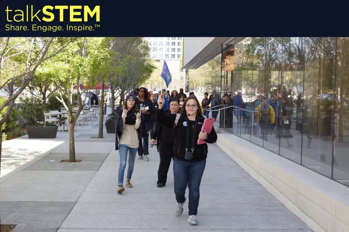 talkSTEM: Share. Engage. Inspire. Photo shows a group of people walking through an urban environment of mostly concrete and glass with a row of young trees alonside. The group's leader is wearing an ear microphone and holding a small blue flag high enough for the group to see and follow.
