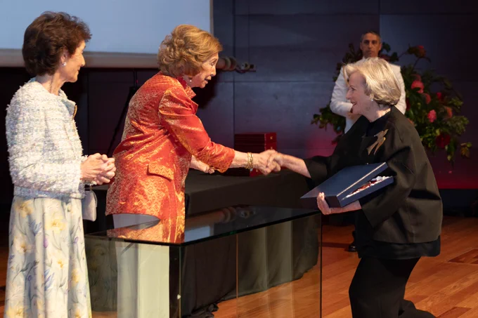En la imagen, Patricia Phelps de Cisneros está recibiendo un premio. Está estrechando la mano de la SM Reina Doña Sofía, quien viste una chaqueta roja con un patrón. Paloma O'Shea, vestida con un vestido floral de colores claros, se encuentra al lado aplaudiendo. El escenario parece ser una ceremonia formal en interiores, con una mesa de vidrio en primer plano y una persona de pie en el fondo, junto a un arreglo floral.