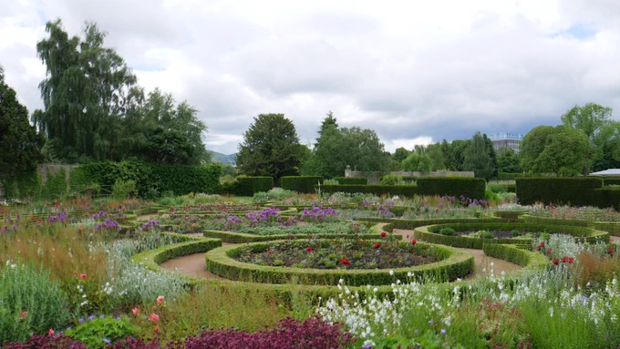 a formal garden with circular beds enclosed by low hedges and containing a profusion of colourful flowers. int the background taller hedges and trees.