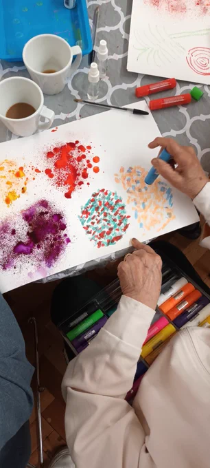 A participants hands making a brightly coloured picture of dotted patterns in vibrant colours. Empty tea cups amd materials on the table. 