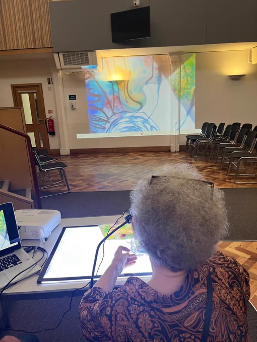 A participant with a light box in a large room with their colourful swirling artwork projected onto the wall.