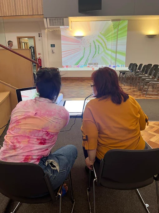 A participant with artist Georgia using a light box in a large room with their colourful swirling artwork projected onto the wall.
