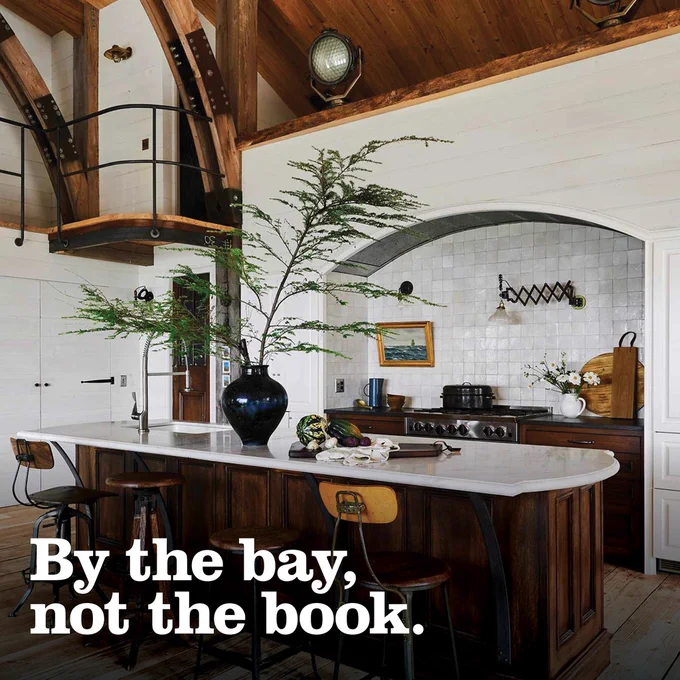 An elegant kitchen with wooden cabinets, exposed wooden beams on the ceiling, and decorative plants on the island countertop. The text "By the bay, not the book." is overlaid on the bottom of the image.