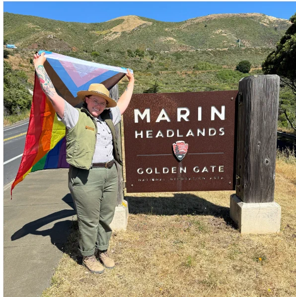 A uniformed NPS Park Ranger stands in front of a sign that says, "Marin Headlands Golden Gate National Recreation Area" while holding a Pride flag