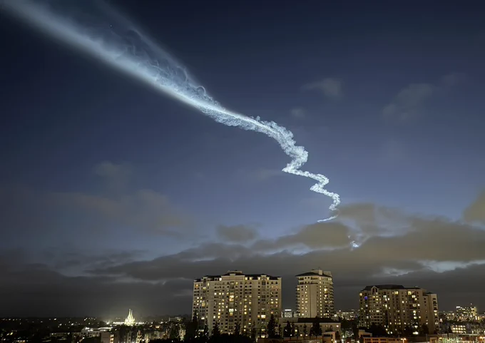 A view of a rocket launch vapor trail in the sky with buildings on the horizon