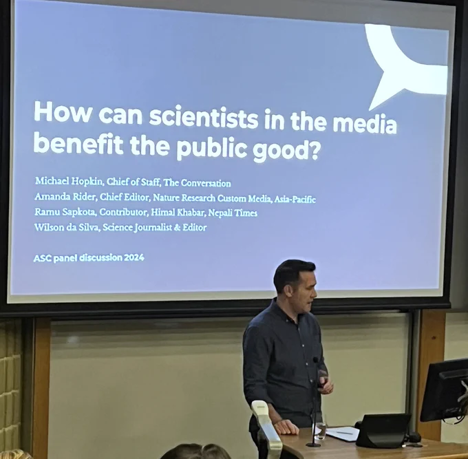 A person stands at a podium in a conference setting, addressing an audience. Behind the speaker, a projected slide reads "How can scientists be in the media" with names and titles of panelists listed: Michael Hopkin, Amanda Rider, Ramu Sapkota, and Wilson da Silva, referring to an upcoming ASC panel discussion in 2024. 
