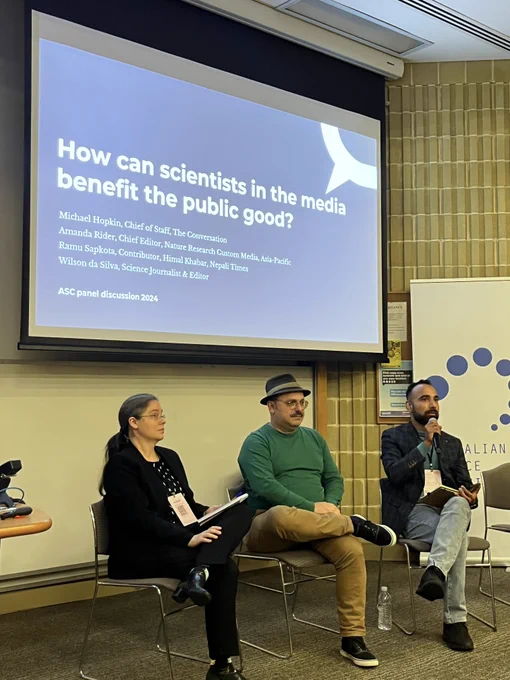 Three panelists are seated in front of an audience at the Australian Science Communicators conference, with a presentation slide displayed behind them titled "How can scientists truly benefit the public good?" featuring names of the speakers and their affiliations.