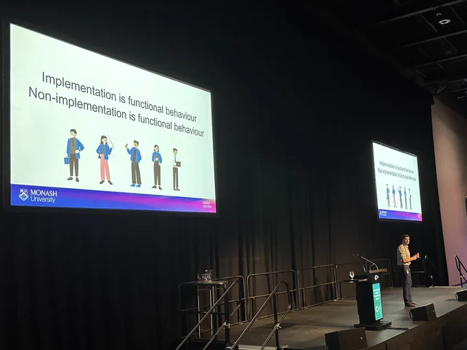 Russell Fox presenting plenary session. Slide displays “Implementation is functional behaviour. Non-implementation is functional behaviour.”