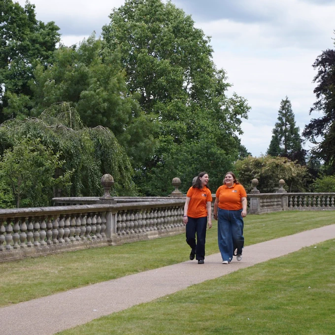 Two student ambassadors in orange tops talking and laughing whilst walking along a path outside Founder's building, surrounded by trees and green spaces.