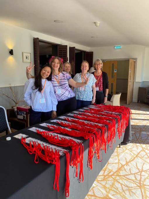 Five women wearing orange lanyards stand behind a table covered in red lanyards and name badges. The room is in a hotel with a patterned carpet. 