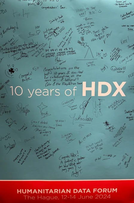 A poster celebrating “10 years of HDX” is covered with handwritten congratulatory messages, doodles, and signatures. The poster background is blue, with “10 years of HDX” prominently displayed in white text. The bottom indicates the Humanitarian Data Forum in The Hague, June 2024.