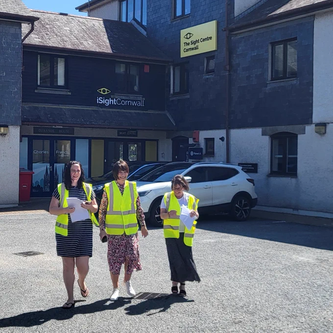 Three women wearing high-vis jackets walk away from a large building with a yellow sign on it that reads iSightCornwall The Sight Centre. In their hands they are holding paperwork