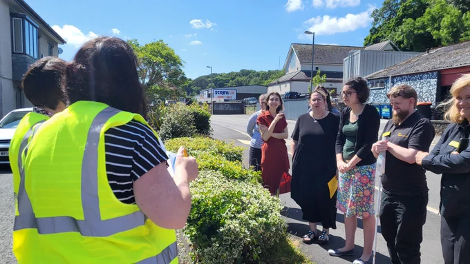 A group of seven people stand outside, facing two women wearing high-vis jackets. Some are wearing office wear whilst two of the people are wearing all black and a yellow iSightCornwall name badge. Behind them is an industrial estate