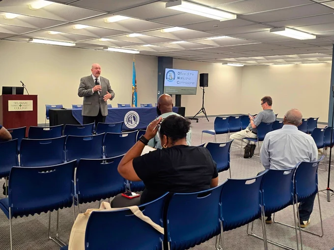 A man standing and speaking with a microphone in a room with empty chairs and a few seated attendees. A screen behind him shows text that reads "Office of the... Commissioner.