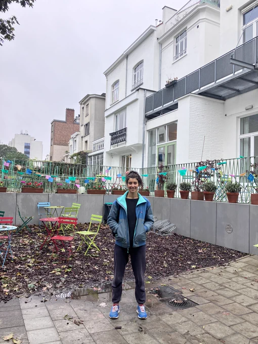 A woman standing outside, next to a white house and colourful chairs