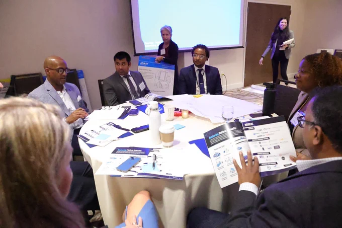 A group of professionals sits around a round table engaged in discussion, with papers and documents spread out. A presenter and another person stand in the background.