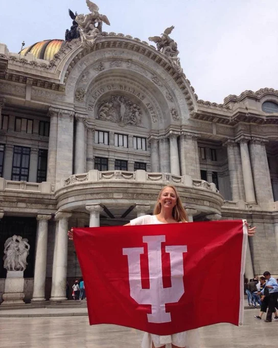 A smiling blonde woman holds a large IU flag as she stands in front of the ornate marble facade of the Palacio de Bellas Artes.