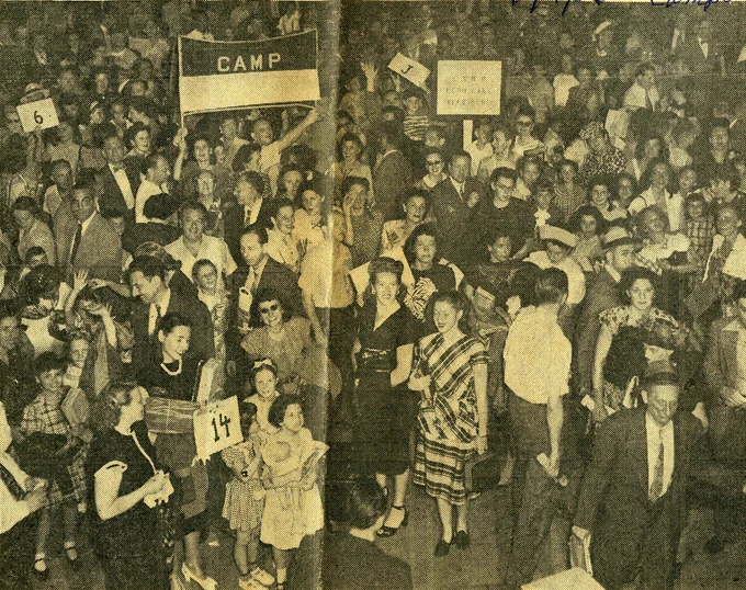 A black and white with yellow tinge photo of a large crowd of young people in period dress. A large sign reading “Camp” can be seen in the crowd being held up by two people. Other smaller handheld signs reading “6,” “J,” and “14” can be seen throughout the crowd.