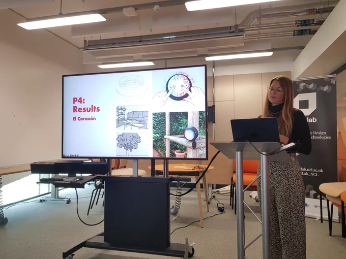 Sara Wolf giving her presentation. The screen says: P4 Results. And has photos of the device they made as part of her research where couples can use it to bring their pulses together. 