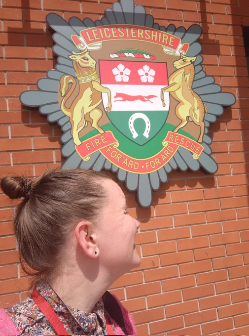 A blonde woman looking away from the camera, squinting, in front of the Leicestershire Fire and Rescue Service crest hanging on a brick wall