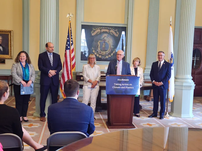 Members of the Massachusetts state Senate stand at the front of a room in the State House behind a podium, an American flag behind them.