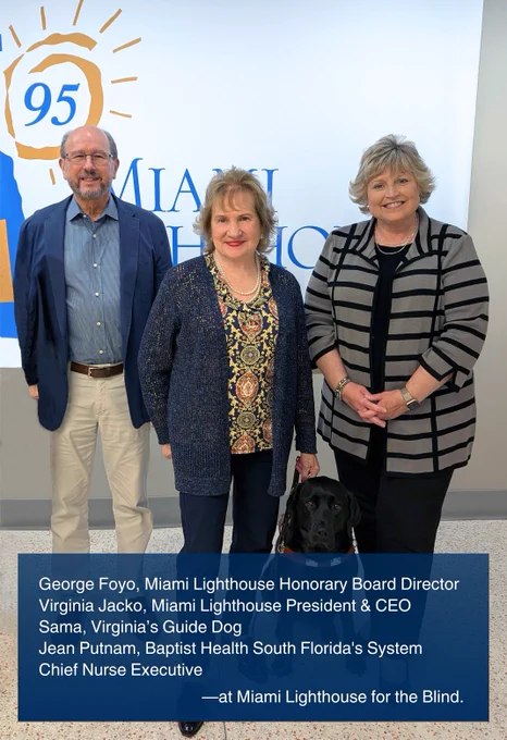 Pictured is George Foyo, Miami Lighthouse Honorary Board Director, Virginia Jacko, Miami Lighthouse President and CEO, holding the leash of her guide dog seated at her feet, and Baptist Health South Florida's System Chief Nurse Executive Jean Putnam. They smile for the camera, standing in front of the Miami Lighthouse for the Blind logo inside the Miami Lighthouse campus.