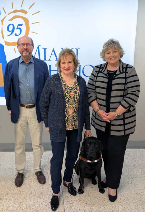 Pictured is George Foyo, Miami Lighthouse Honorary Board Director, Virginia Jacko, Miami Lighthouse President and CEO, holding the leash of her guide dog seated at her feet, and Baptist Health South Florida's System Chief Nurse Executive Jean Putnam. They smile for the camera, standing in front of the Miami Lighthouse for the Blind logo inside the Miami Lighthouse campus.