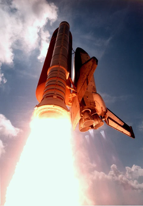 A Space Shuttle, identifiable by its design and markings, launches into a clear sky, exhibiting a powerful ascent with fiery rocket boosters.