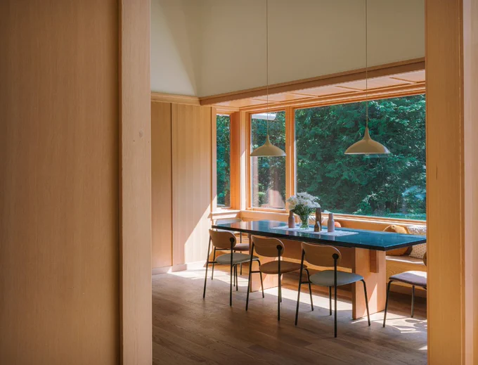 Modern dining room with a large wooden table, black chairs, and pendant lights, overlooking a lush green forest through floor-to-ceiling windows.
