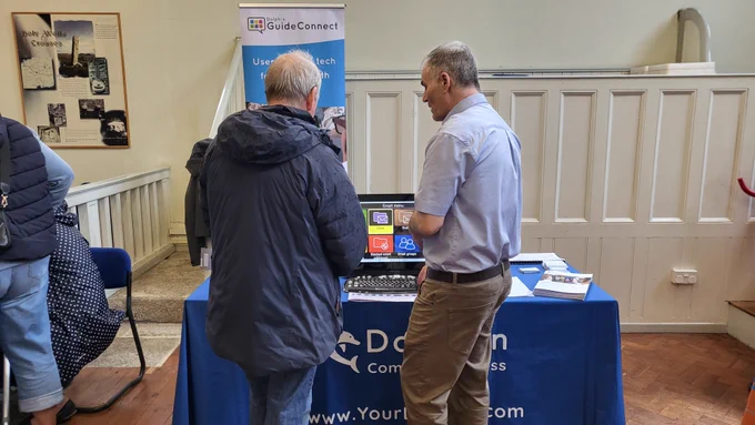 Two men stood in front of a table. The table has a display on it of GuideConnect, software for people who are visually impaired or blind. One man is in a shirt demonstrating to the other man.
