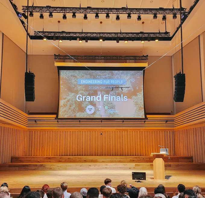 Wooden panelled hall with an empty stage. A projector screen shows a slide saying EWB Grand Finals.