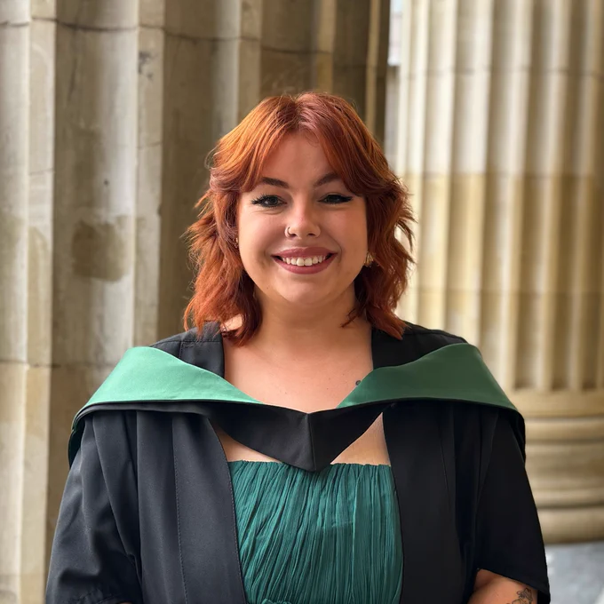 Wimberley Award winner Dani posed in front of the Caird Hall pillars on her graduation day, she is smiling and wearing a black graduation gown and and a graduation hood with green lining 