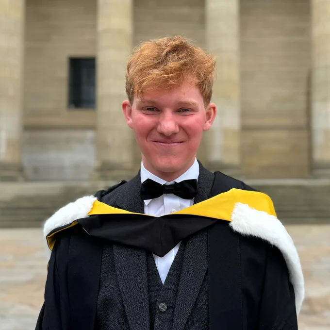 Wimberley Award winner Kai posed in front of the Caird Hall pillars on his graduation day, he is smiling and wearing a black graduation gown and and a graduation hood with yellow lining and fur on the outer hood