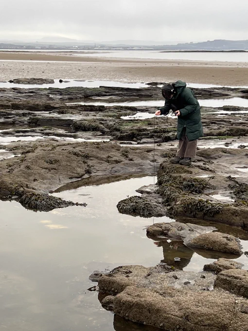 Seascape rockpool scene with figure holding a wire into the water, wearing a long green jacket and headphones