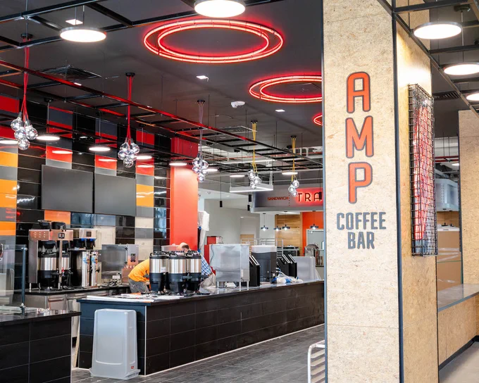 A dining venue with red overhead lights, a red, yellow, and black backsplash, and the words "AMP Coffee Bar" on a pole. 