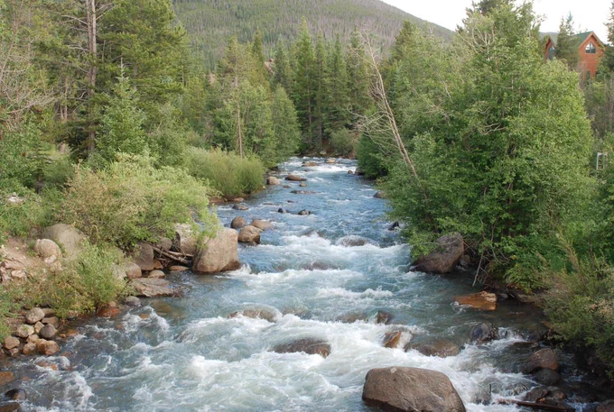 Photograph of the Snake River, Rocky Mountains, at 9,000 feet. We see a fast-flowing river with large rocks. The riverbanks are lined with trees. 
