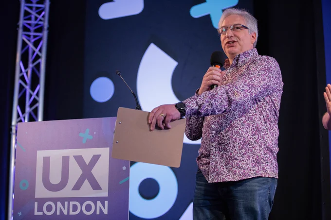 A man is on stage talking in to a handheld microphone. He is gesturing with a clipboard in the other hand. Behind him large graphic shape decorations and to his left is a UX London branded podium.