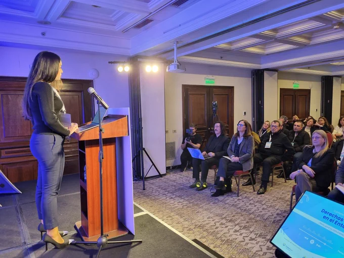 Ministra Facio, entre el público, escucha a Vila, que habla desde un escenario.
