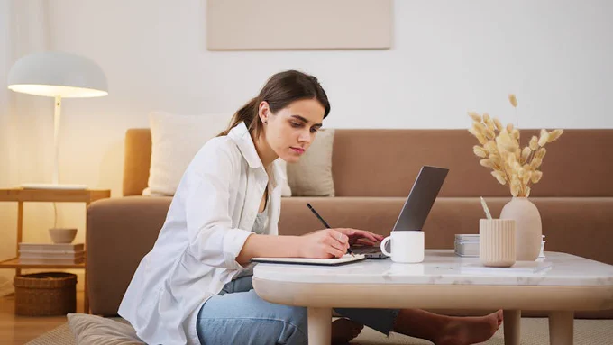 Side view full length focused female in casual clothes browsing laptop and writing down notes while sitting on floor in cozy modern apartment.