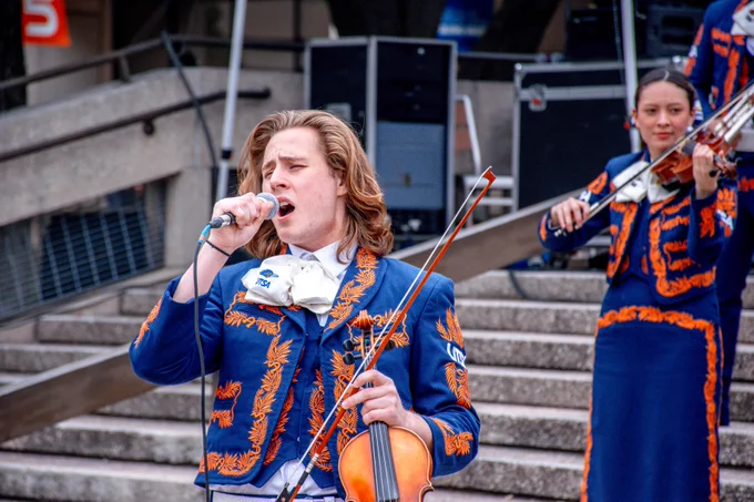 A musician in a blue and orange patterned suit performs passionately into a microphone, holding a violin, with another musician in a matching outfit playing a violin in the background.