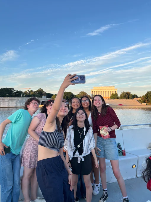 Summer Academy fellows pose for a selfie on a boat. In the background the sun is setting over the Lincoln Memorial.