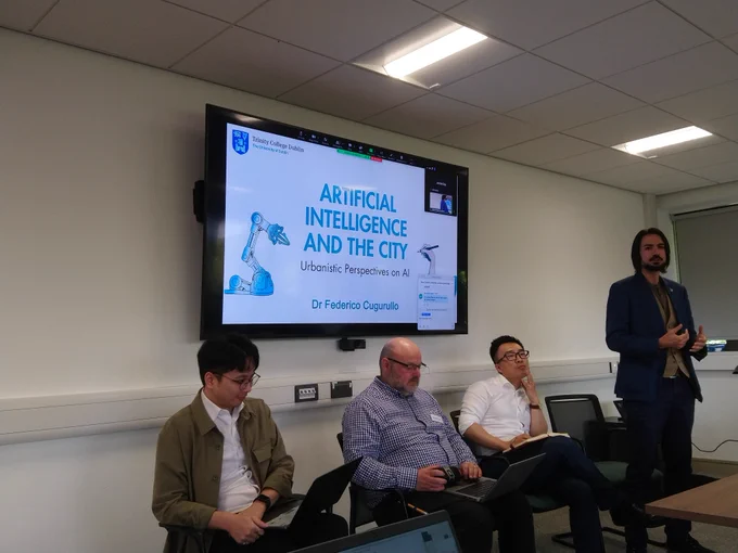 Image of the book panel. Three people sitting and one person standing. Behind them is a screen showing a slide show with the words Artifical Intelligence and the City :urbanistic perspectives on AI
