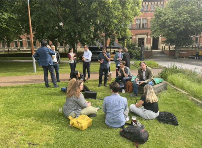 Image of a group of 13 people standing and sitting on grass in the sunshine