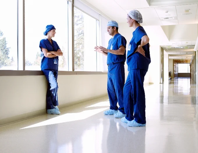 Three surgeons in scrubs stand in a brightly lit hospital hallway talking.