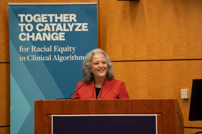 Helen Burstin at the podium. A blue banner with the text "Together to Catalyze Change for Racial Equity in Clinical Algorithms" appears in the background. 