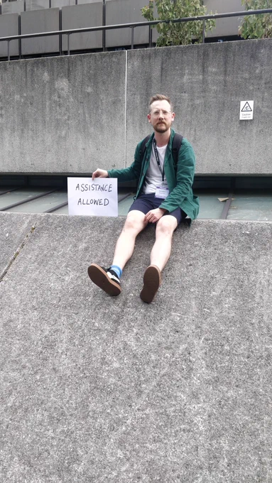 A man sitting on a wall holding a sign saying assistance allowed, behind him a sign days caution fragile roof