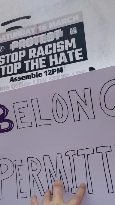 A hand written sign saying belonging permitted held up to a poster saying stop racism, stop the hate, assemble 12pm