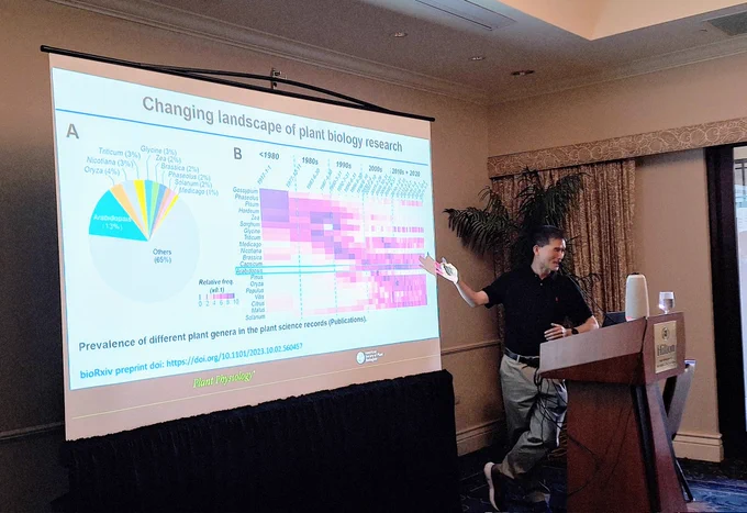 A person standing behind a podium pointing to a screen behind them. The screen shows two colorful charts of data under a title that says "Changing landscape of Plant Biology Research"