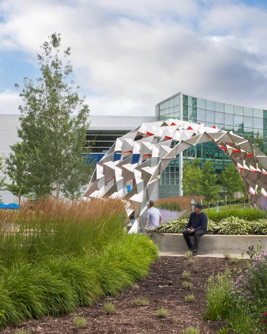 A person sitting outdoors with an arch-shaped structure behind them and a glass-paneled building in the distance.